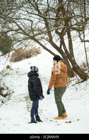 Ein Mann und ein Kind, warm in Winterkleidung gekleidet, stehen in einer schneebedeckten Landschaft. Das Kind trägt einen kleinen Schlitten. Der Schnee fällt sanft und erzeugt eine Erbse Stockfoto