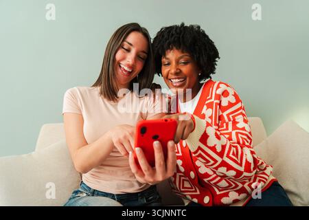 Zwei glückliche Frauen sitzen eng zusammen, während sie ein Smartphone verwenden, um spannende Anwendungen anzusehen und spannende Momente zu teilen und ihre zu zeigen Stockfoto