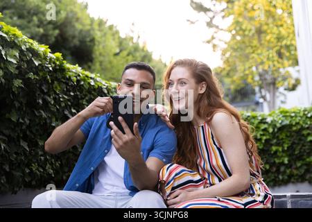 Junges Paar sitzt draußen, lächelt und nimmt Selfie mit dem Smartphone Stockfoto