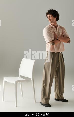 Der junge Mann zeigt sein Gespür für Mode und trägt ein schickes Outfit, während er neben einem Stuhl steht. Stockfoto