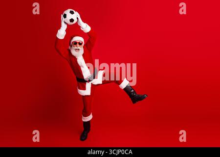 Der Weihnachtsmann trägt eine Sonnenbrille und spielt Fußball, hält einen Ball vor rotem Hintergrund für Weihnachtsspaß und -Freude Stockfoto