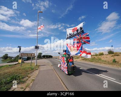 Sheerness, Kent, Großbritannien. September 2025. Der 80-jährige englische Exzentriker Tim Bell sieht heute eine Reihe von Englands Nationalflaggen (das St. George's Cross) entlang der Küstenstraße in Sheerness, Kent, zusammen mit seinem eigenen angeflaggten, maßgeschneiderten Roller „Tank“. Mr. Bell (von Minster on Sea) nutzt seine beiden umgebauten Mobilitätsroller, um Geld für den Kent Surrey Sussex Luftwagenwagen zu sammeln. Die Operation Raise the Colours hat die Öffentlichkeit dazu ermutigt, Englands Nationalflaggen zu fliegen. Quelle: James Bell/Alamy Live News Stockfoto