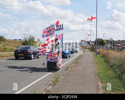 Sheerness, Kent, Großbritannien. September 2025. Der 80-jährige englische Exzentriker Tim Bell sieht heute eine Reihe von Englands Nationalflaggen (das St. George's Cross) entlang der Küstenstraße in Sheerness, Kent, zusammen mit seinem eigenen angeflaggten, maßgeschneiderten Roller „Tank“. Mr. Bell (von Minster on Sea) nutzt seine beiden umgebauten Mobilitätsroller, um Geld für den Kent Surrey Sussex Luftwagenwagen zu sammeln. Die Operation Raise the Colours hat die Öffentlichkeit dazu ermutigt, Englands Nationalflaggen zu fliegen. Quelle: James Bell/Alamy Live News Stockfoto