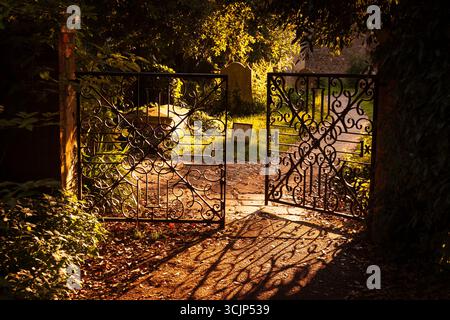 Am Abend Sonnenlicht wirft seinen Schatten Muster der teils offenen schmiedeeisernen Tor über den Weg zur Bosham Kirchhof. Stockfoto