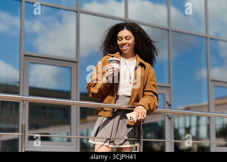 Eine stilvolle junge Frau mit lockigen Haaren isst ihren Kaffee, während sie für den Herbst in einem schicken Outfit gekleidet ist. Stockfoto