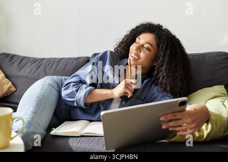 Die Frau mit lockigen Haaren entspannt sich auf der Couch, während sie zu Hause freischaffende Aufgaben wahrnimmt. Stockfoto