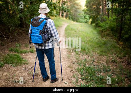 Senior man wandert auf einem bergigen Waldweg mit Rucksack und Wanderstöcken. Stockfoto