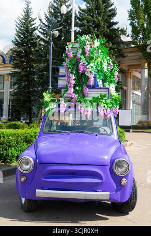 Ein altes lila Auto mit üppigen grünen und lila Blumen und einem Stapel Bücher auf dem Dach, das in einem Park geparkt ist Stockfoto