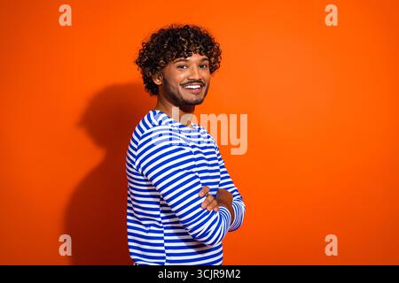 Junger Mann in einem gestreiften, von Seemännern inspirierten Hemd posiert vor einem orangen Hintergrund mit einer fröhlichen und selbstbewussten Einstellung Stockfoto