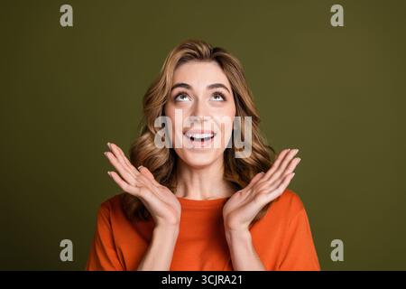 Junge, fröhliche Frau im orangefarbenen T-Shirt lächelt, während sie vor khakifarbenem Hintergrund mit erhobenen Händen Freude ausdrückt Stockfoto