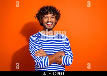 Fröhlicher junger Mann in einem gestreiften Hemd, der die Arme vor einem orangefarbenen Hintergrund überkreuzt, in einem legeren und stilvollen Look Stockfoto