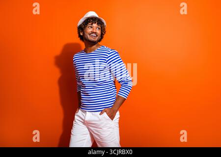 Positiver, stylischer Mann im gestreiften Matrosen-Outfit, der vor leuchtend orangefarbenem Hintergrund posiert und Sommermode und Reisen repräsentiert Stockfoto