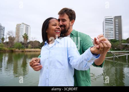 Glückliches multirassisches Paar, das am See im Stadtpark tanzt. Stockfoto