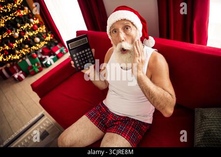 Der Weihnachtsmann hält einen Taschenrechner in einem festlichen Wohnzimmer mit Weihnachtsdekoration und sieht überrascht aus, während er lässige Urlaubskleidung trägt Stockfoto