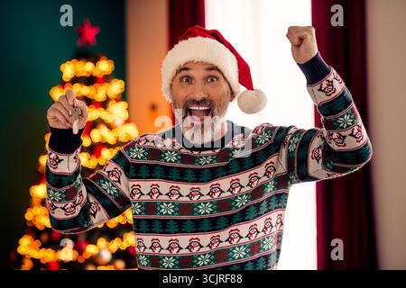 Fröhlicher älterer Mann mit Schlüsseln in einem festlich gestalteten Raum mit Weihnachtsbaum und lebhaftem Weihnachtsleben Stockfoto