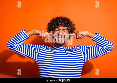 Junger Mann in gestreiftem Hemd posiert spielerisch vor orangem Hintergrund, bedeckt Ohren mit Fingern und lächelt weit Stockfoto