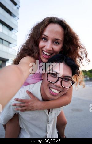 Selfie eines jungen Paares, das in die Kamera schaut, während der Freund seiner Freundin Huckepack gibt. Stockfoto