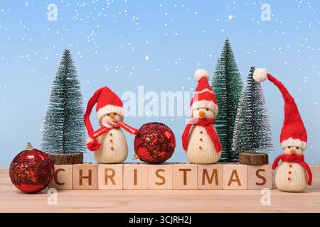 Weihnachtshintergrund mit gestrickten Schneemännern in roten Hüten und Weihnachtsbäumen mit roter Kugel und Holzwürfeln mit dem Wort Weihnachten auf ihnen. Weihnachten ca. Stockfoto