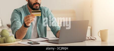 Araber, der online mit Laptop und Kreditkarte bezahlt, zu Hause in Kithcen sitzt, leerer Raum Stockfoto