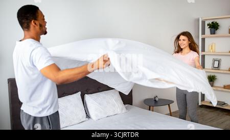 Hausarbeiten. Millennial multinationales Paar in der Liebe macht Bett zu Hause, Spaß haben, während Hausarbeit zusammen Stockfoto