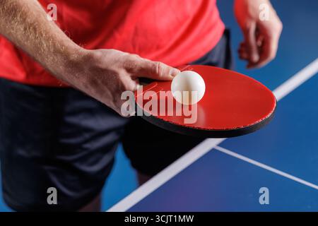 Nahaufnahme eines Spielers, der eine Tischtennisplatte mit rotem Paddel und Ball auf einem blauen Tisch vorbereitet, Indoor Training und Wettkampfkonzept Stockfoto
