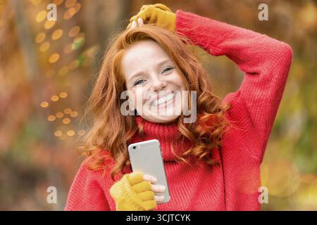 Rothaarige fröhliche Frau im Herbst mit Smartphone in der Hand, Porträt Stockfoto