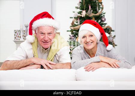 Seniorenpaar mit Weihnachtsmannmützen auf einem weißen Sofa, einem Weihnachtsbaum im Hintergrund Stockfoto