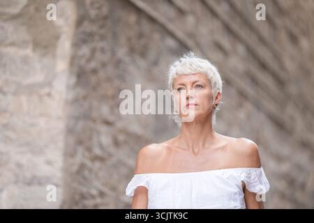 Eine Frau in einem weißen Kleid steht vor einer Steinmauer. Sie hat einen ernsthaften Ausdruck im Gesicht Stockfoto