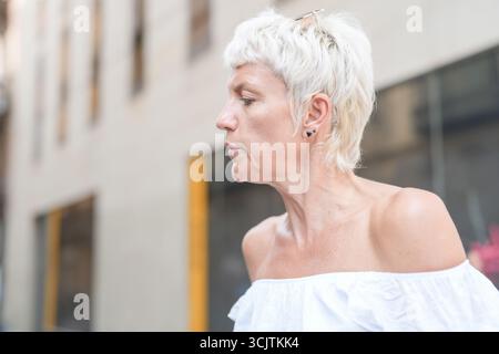 Eine Frau mit kurzen blonden Haaren und einem weißen Hemd steht vor einem Gebäude. Sie hat einen ernsthaften Ausdruck im Gesicht Stockfoto