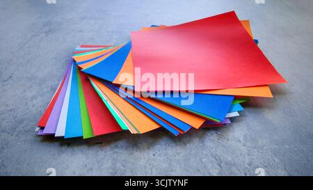 Ansicht von oben nach unten eines Stapels aus mehrfarbigem Origami-Papier auf einem grauen Stein- oder Betonhintergrund. Stockfoto