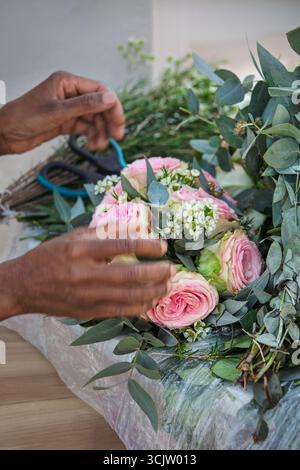 Florist im Blumengeschäft, der Blumenarrangements zusammenstellt, einen Brautstrauß mit rosa Rosen macht, afrikanische Frauenhände mit einer Schere Stockfoto