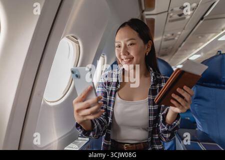 Eine junge Frau sitzt auf einem Flugzeugsitz, lächelt, während sie ihr Handy und ihren Pass hält, Selfies macht und den Flug genießt. Sie sieht entspannt aus und Stockfoto