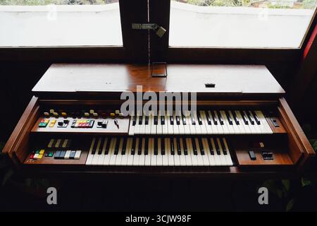 Elektrische Retro-hölzerne Orgel mit mehreren Schlüsseln sitzt drinnen in der Nähe eines Fensters und bietet eine klassische Musikinstrumentenszene für Retro-Fans. Stockfoto