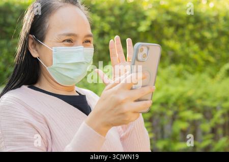 Frau mit Schutzmaske. Glückliche asiatische Frau, die eine medizinische Maske trägt und ein Telefon im Freien im Naturpark benutzt. Stockfoto