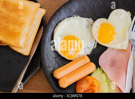 Ein köstliches Frühstück mit herzförmigem Spiegelei, Schinken, Wurst und Toast. Frühstückskonzept. Stockfoto