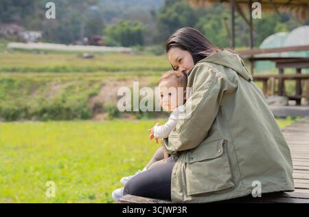 Glückliche asiatische Mutter umarmt ihr Baby, während sie bei Sonnenuntergang im natürlichen Garten sitzt. Familie verbringt Zeit zusammen im Freien. Stockfoto