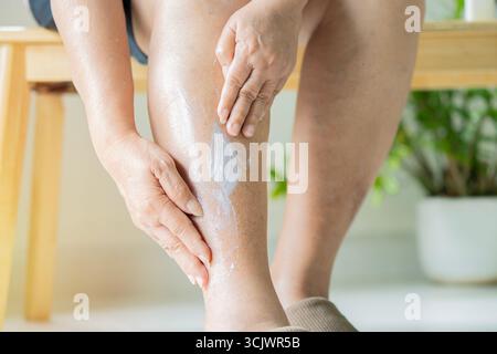 Nahaufnahme einer älteren Frau, die Lotion aufwendet, während sie auf einem Holzstuhl im Wohnzimmer sitzt. Senior Healthy Konzept. Stockfoto