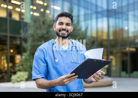 Ein zuversichtlicher und freundlicher indischer Arzt steht vor einem modernen Krankenhausgebäude, hält ein Klemmbrett mit Patientenakten und lächelt Stockfoto