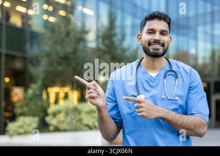Junger männlicher Arzt mit blauen Peelings und Stethoskop zeigt mit beiden Zeigefingern in verschiedene Richtungen vor einem modernen Krankenhausgebäude, lächelt und blickt in die Kamera Stockfoto