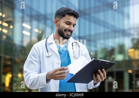 Selbstbewusster junger männlicher Arzt, der Patientenakten außerhalb eines modernen Krankenhauses überprüft, Gesundheitskompetenz und Professionalität verkörpert, während er sich mit Patientenversorgung und -Analyse befasst Stockfoto