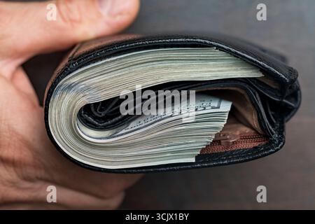 Die Hand eines Mannes hält eine schwarze Ledertasche mit einem Haufen 100-Dollar-Scheine, Banknoten, Bargeld, Nahaufnahme. Konzept für verlorenes oder gefundenes Wallet mit CA Stockfoto