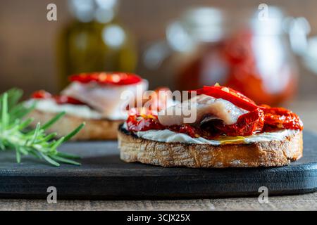 Zwei Brotscheiben mit Ricotta, Heringsstücken und sonnengetrockneten roten Tomaten auf Holzbrett Hintergrund. Köstliches Frühstück oder Wein-Snack. Clos Stockfoto