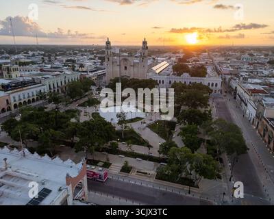 Aus der Vogelperspektive auf die majestätische Kathedrale von Mérida, die einen langen Schatten über der pulsierenden Plaza Grande wirft, die in das warme Licht der untergehenden Sonne getaucht ist, Stockfoto