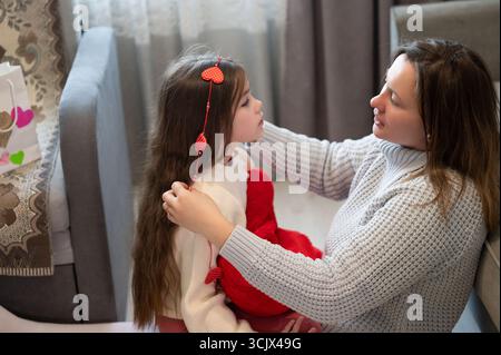 Mutter und Tochter teilen einen ruhigen Moment. Die Mutter passt sanft eine herzförmige Haardekoration im Haar ihrer Tochter an. Die Szene ist warm und intakt Stockfoto