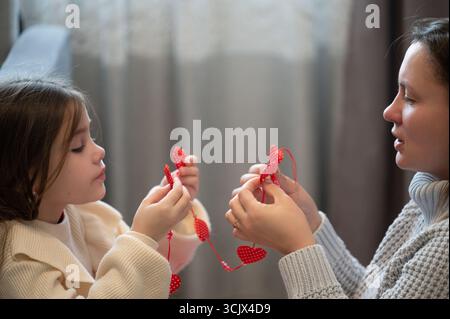 Eine herzerwärmende Szene von Mutter und Kind, die zusammen eine Kette dekorativer roter Herzen zusammenbauen. Sie sitzen in der Nähe und genießen eine gemeinsame Klimaanlage Stockfoto