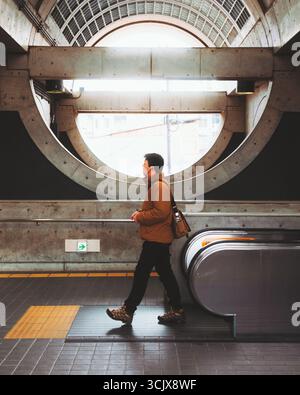 Uji, Japan - 21. Februar 2025: Der Blick auf eine Einzelfigur in einer braunen Jacke geht an der Rolltreppe der Uji Station vorbei, eingerahmt von dem einzigartigen runden Fenster der Station. Stockfoto