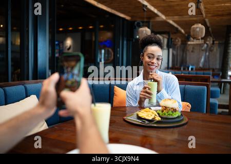 Ein vielseitiges Paar mit Feinschmecker genießt ein zwangloses Abendessen und einen Telefonmoment, bei dem sonnige Vibes und Drinks geteilt werden Stockfoto