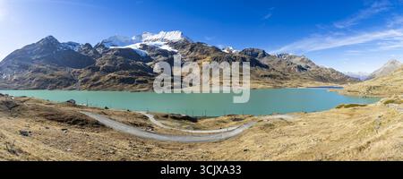 Blick auf das pulsierende türkisfarbene Wasser, das die schneebedeckten Berge unter einem klaren azurblauen Himmel spiegelt, mit gewundenen Pfaden, die durch goldene Felder führen, Samedan Stockfoto