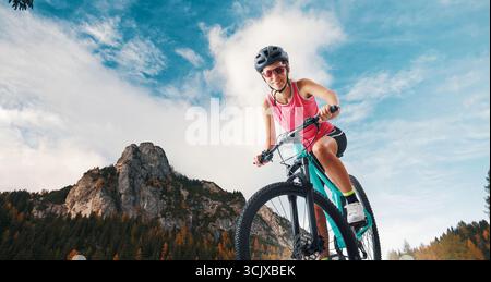 Eine fröhliche junge Frau in Sportkleidung fährt mit dem Mountainbike auf einem Bergweg und lächelt vor einer atemberaubenden Naturkulisse in die Kamera. Perfec Stockfoto