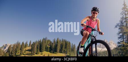 Eine fröhliche junge Frau in Sportkleidung fährt mit dem Mountainbike auf einem Bergweg und lächelt vor einer atemberaubenden Naturkulisse in die Kamera. Perfec Stockfoto